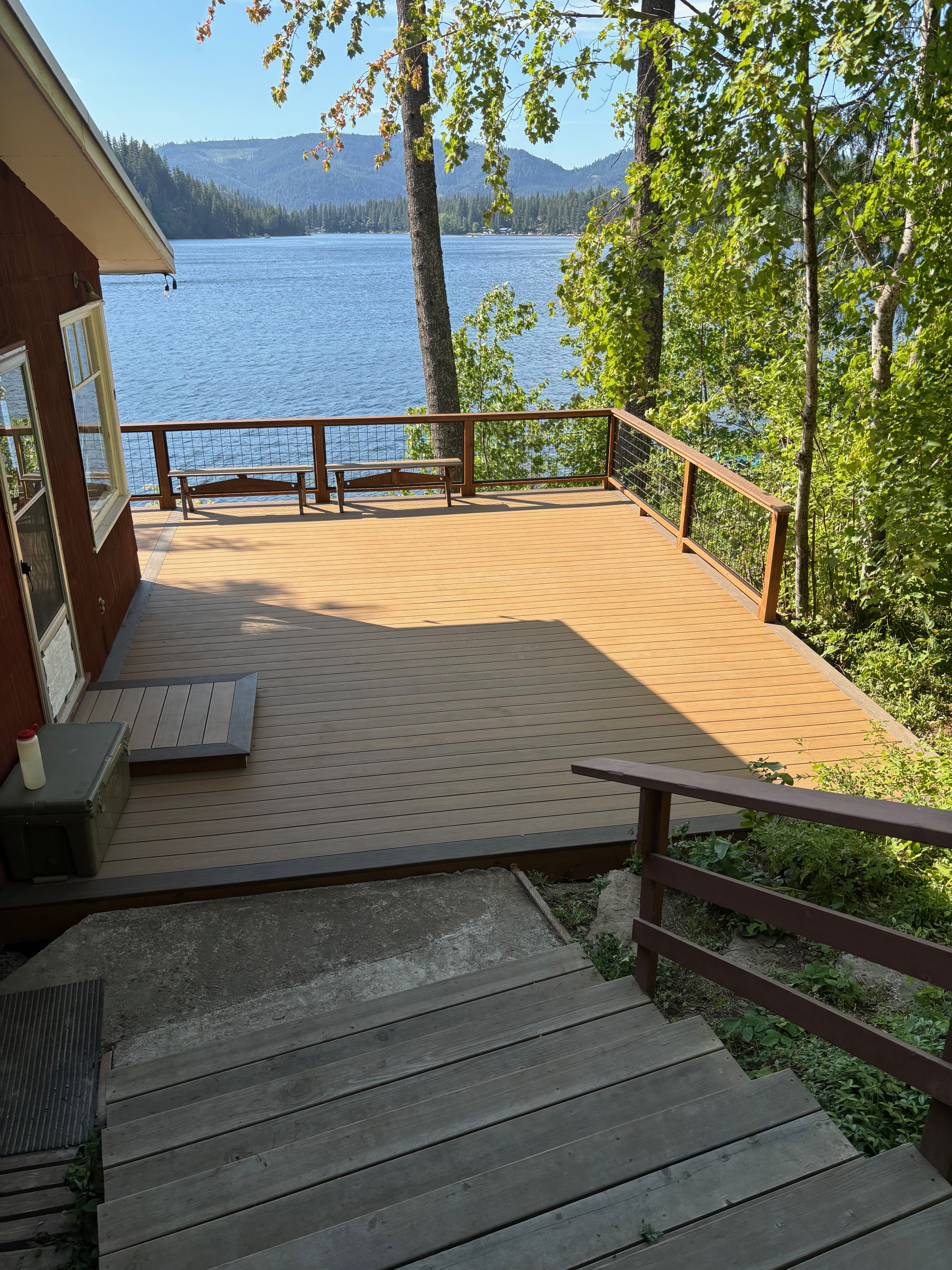 Lakeside composite deck with tan boards, wire mesh railings, and steps down from a forest cabin.