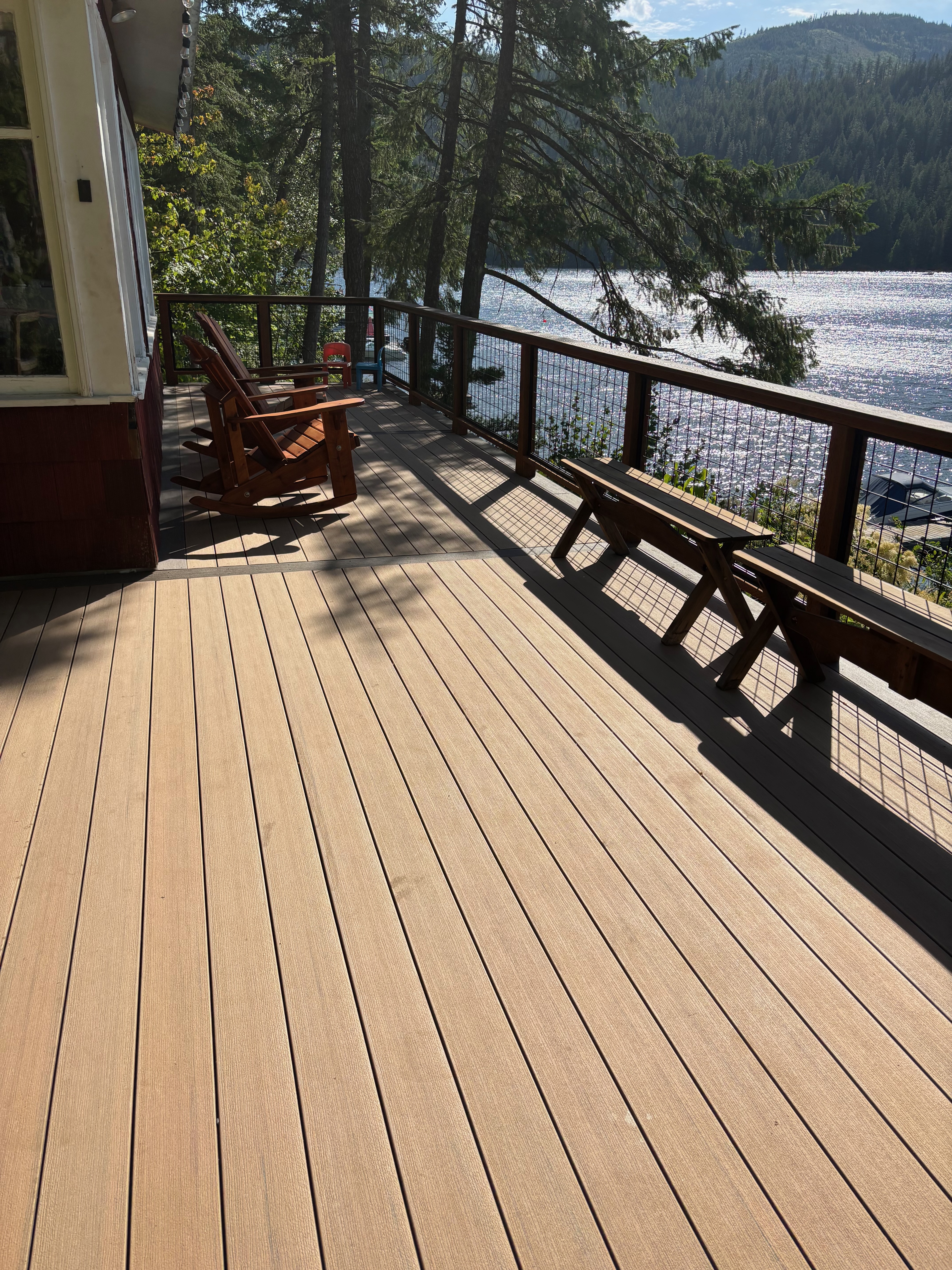 Waterfront deck with built-in benches and wire mesh railing, framed by trees and mountains in the distance.